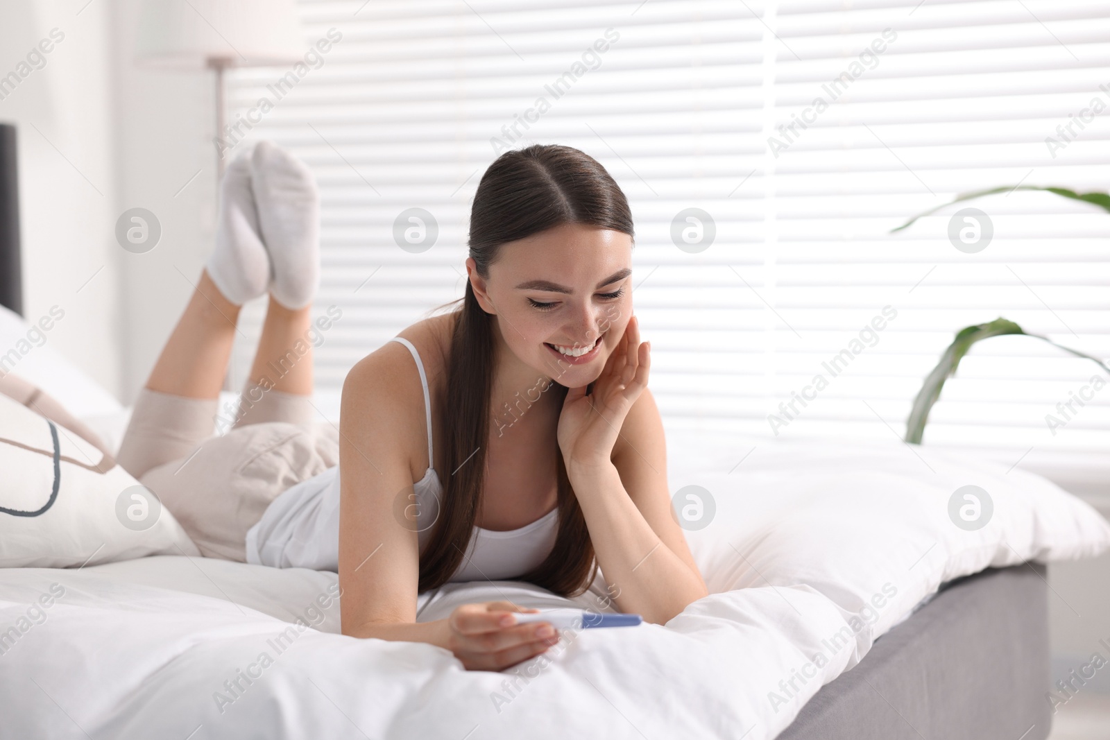 Photo of Happy woman holding pregnancy test on bed in room