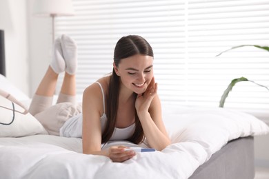 Happy woman holding pregnancy test on bed in room