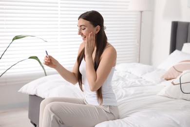 Photo of Happy woman holding pregnancy test on bed in room