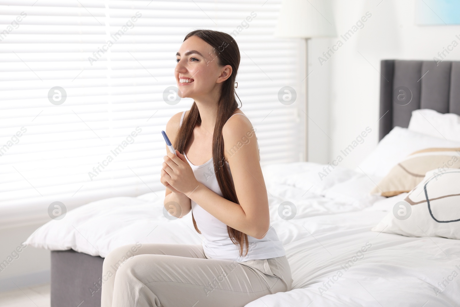 Photo of Happy woman holding pregnancy test on bed in room