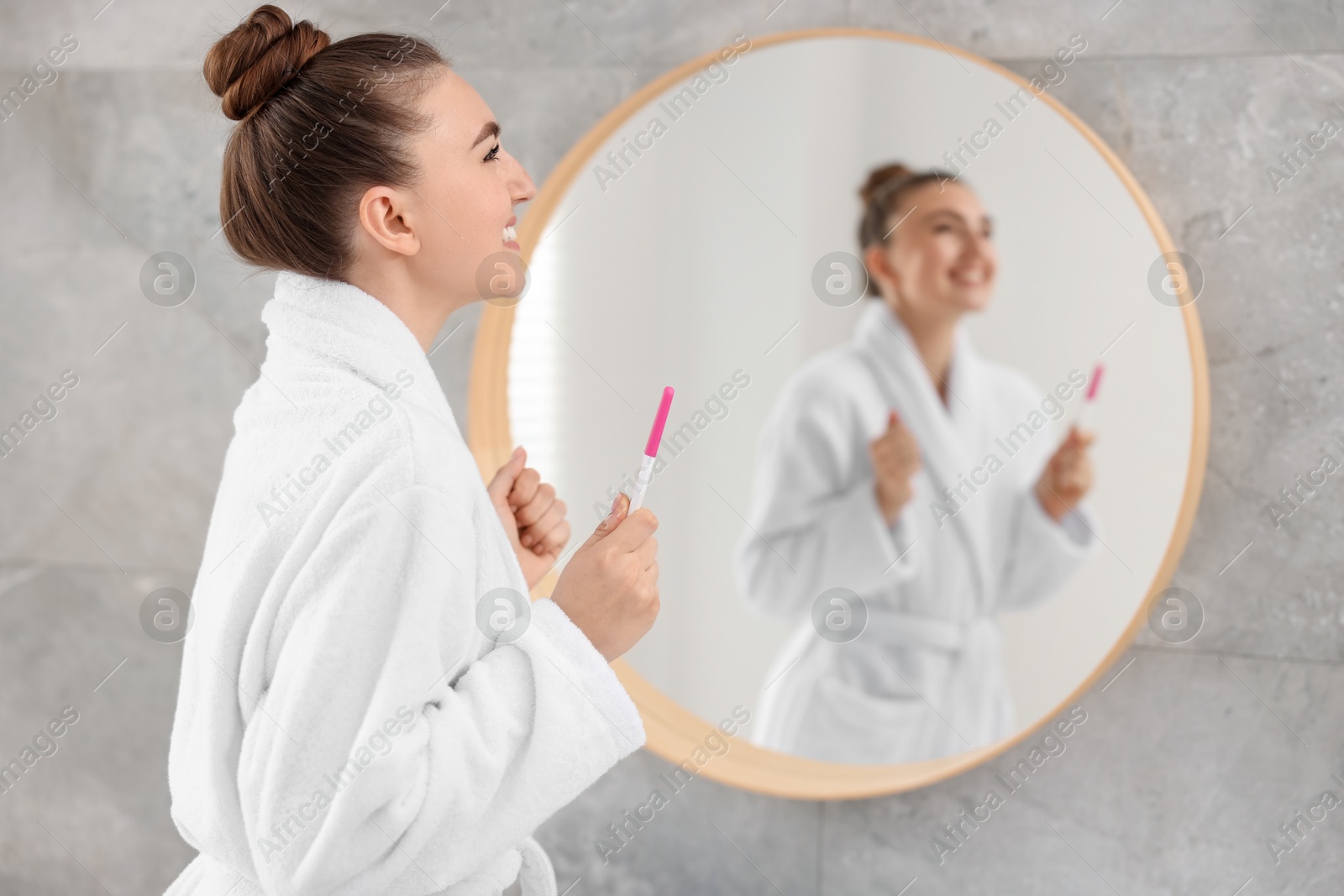 Photo of Happy woman holding pregnancy test near mirror in bathroom