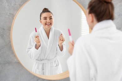 Happy woman holding pregnancy test near mirror in bathroom