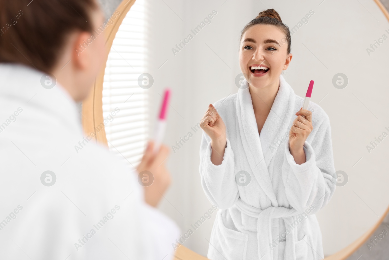 Photo of Happy woman holding pregnancy test near mirror in bathroom