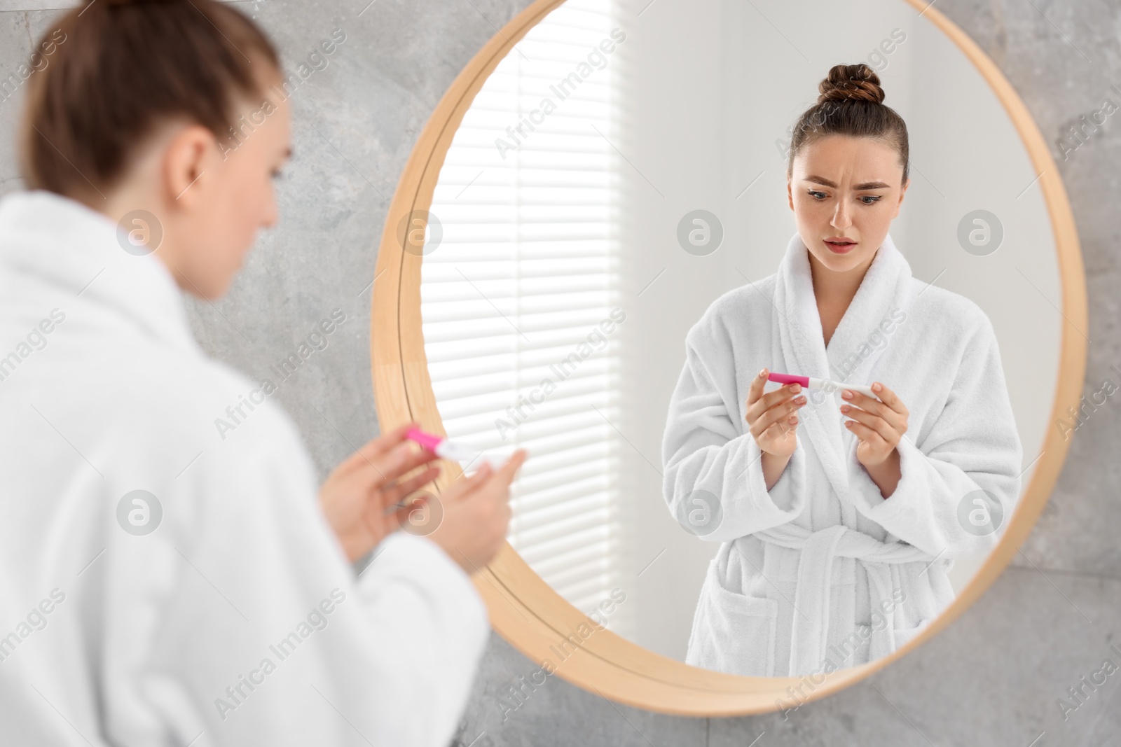 Photo of Sad woman holding pregnancy test near mirror in bathroom