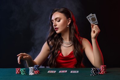 Charming woman playing poker at table on black background with smoke