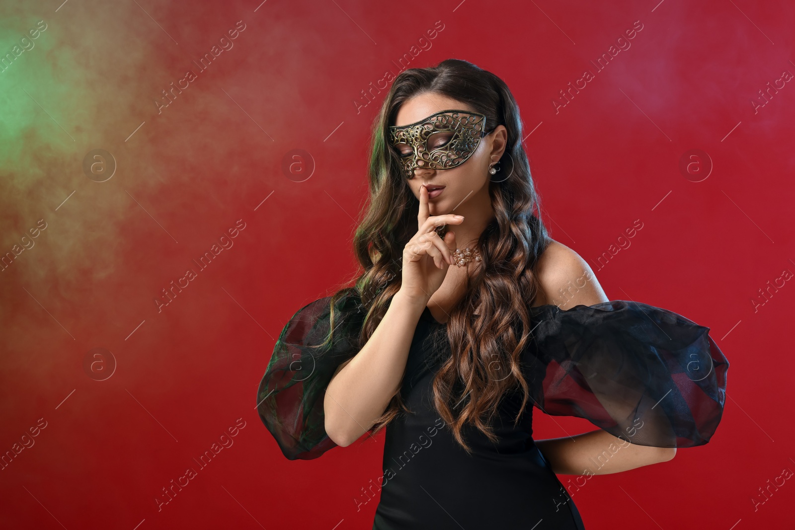 Photo of Beautiful woman with carnival mask showing hush gesture on red background in color lights and smoke. Space for text