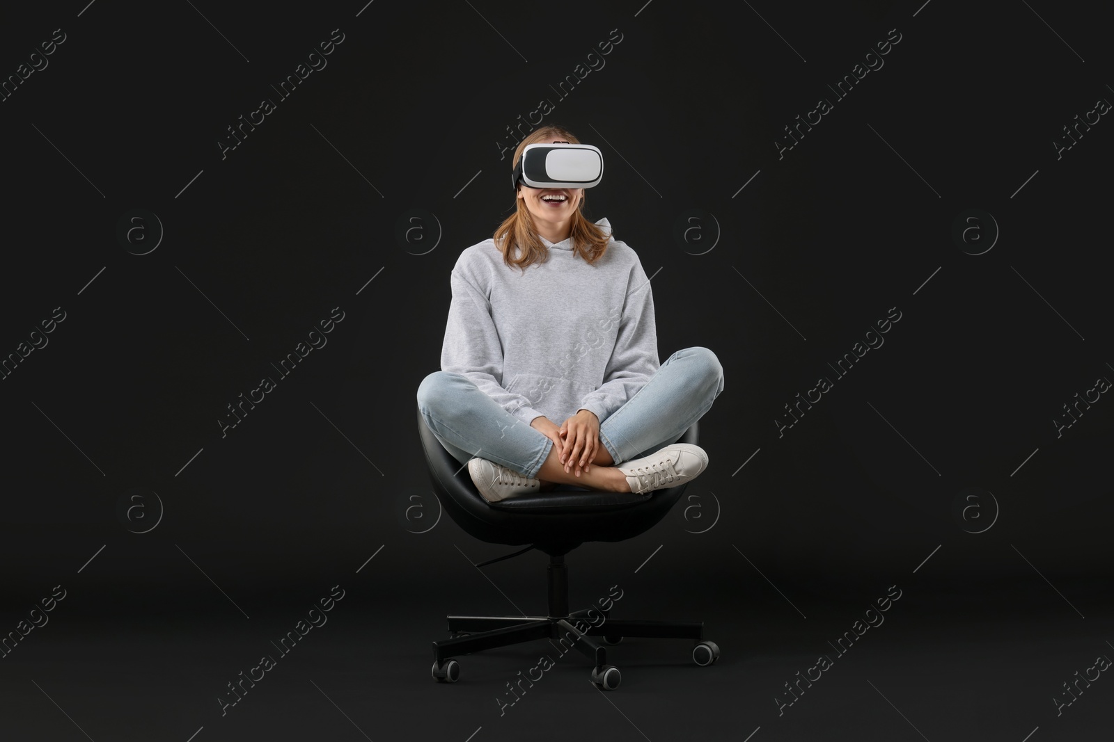 Photo of Happy woman with virtual reality headset sitting on chair against black background