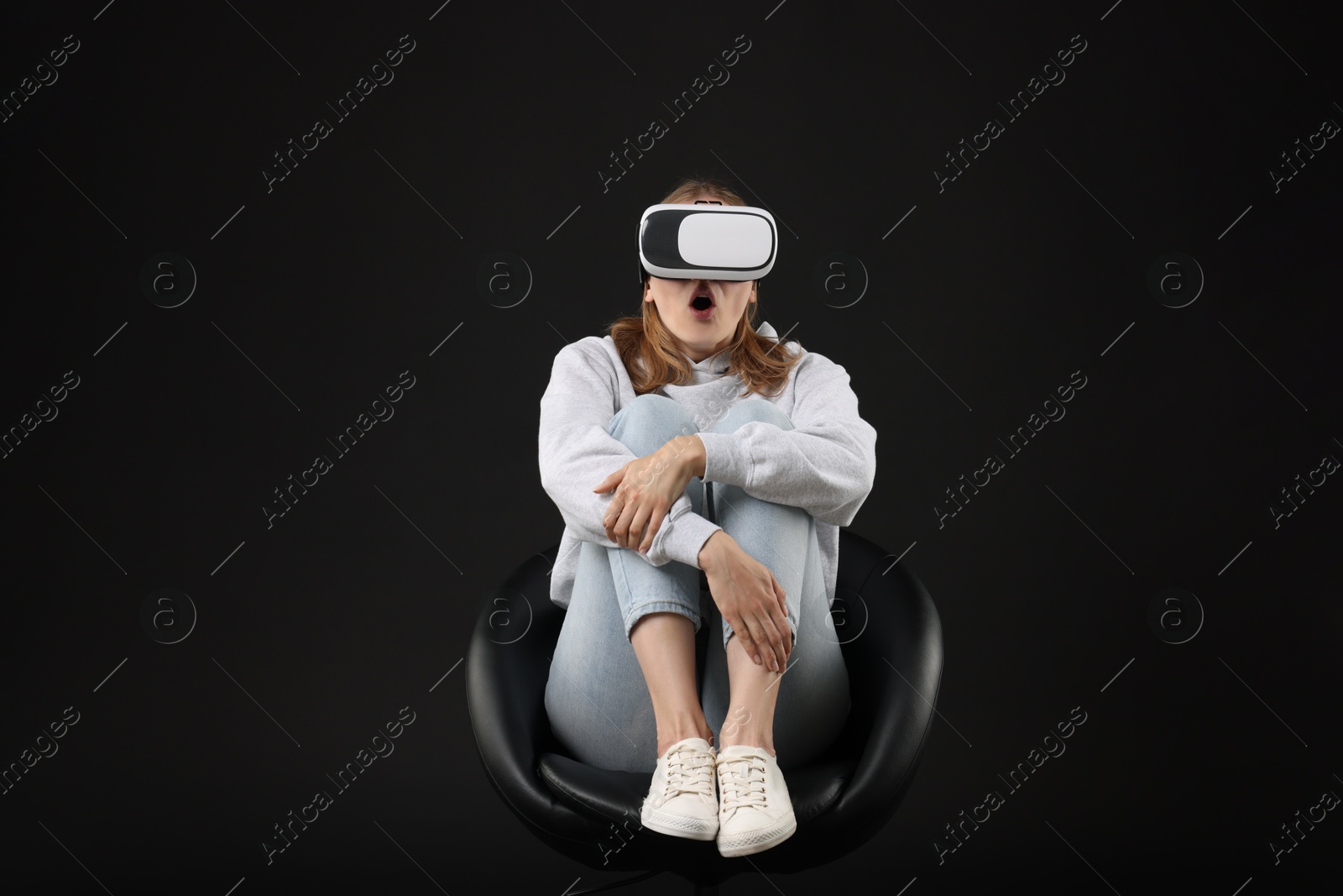 Photo of Emotional woman with virtual reality headset sitting on chair against black background