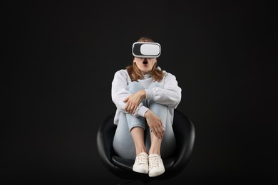 Emotional woman with virtual reality headset sitting on chair against black background