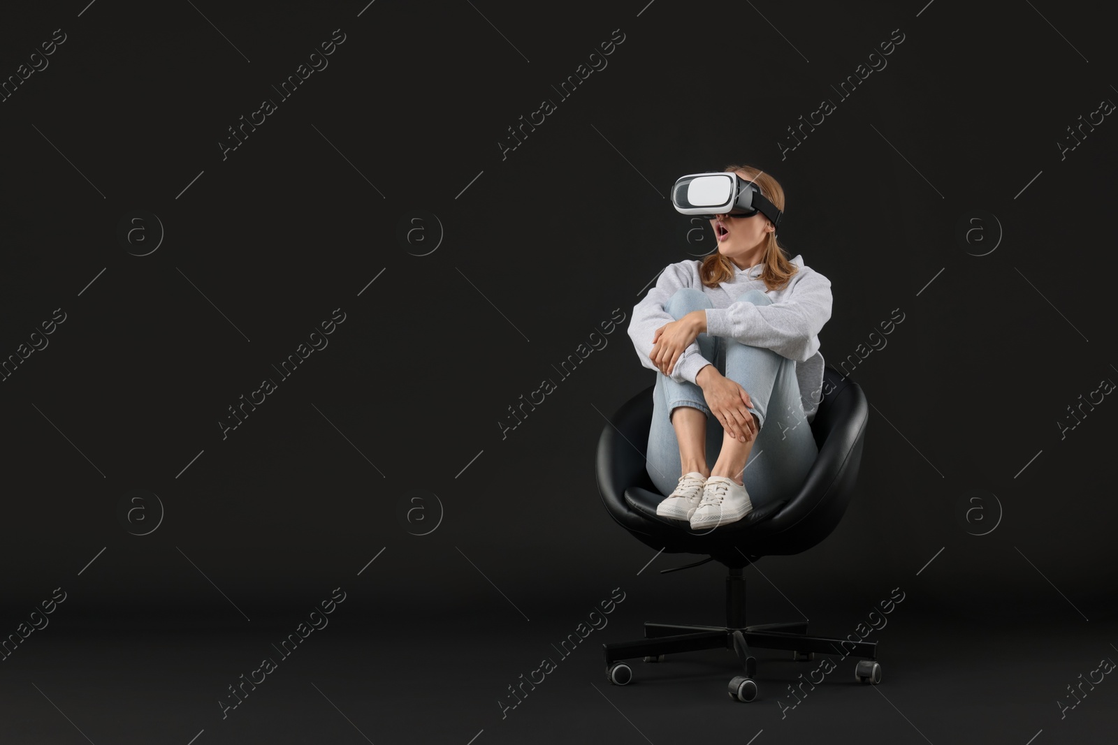 Photo of Emotional woman with virtual reality headset sitting on chair against black background, space for text