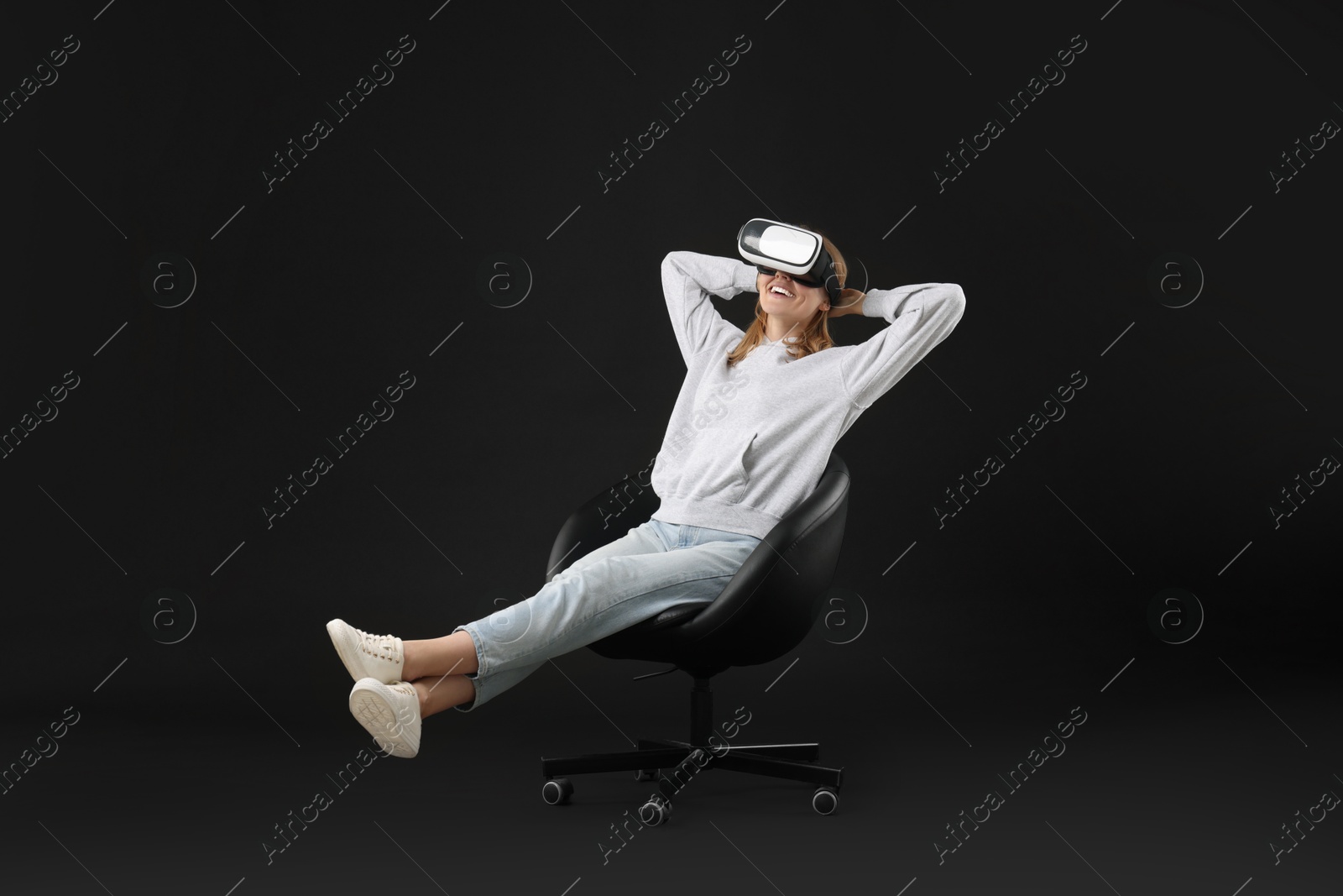 Photo of Happy woman with virtual reality headset sitting on chair against black background