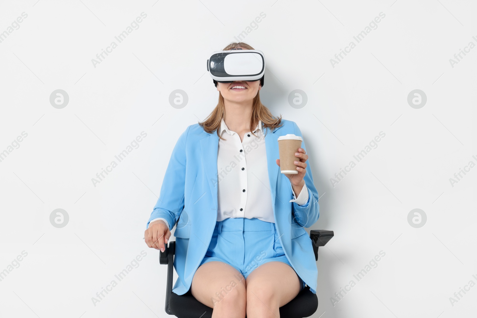 Photo of Happy woman with virtual reality headset and paper cup sitting on chair against white background