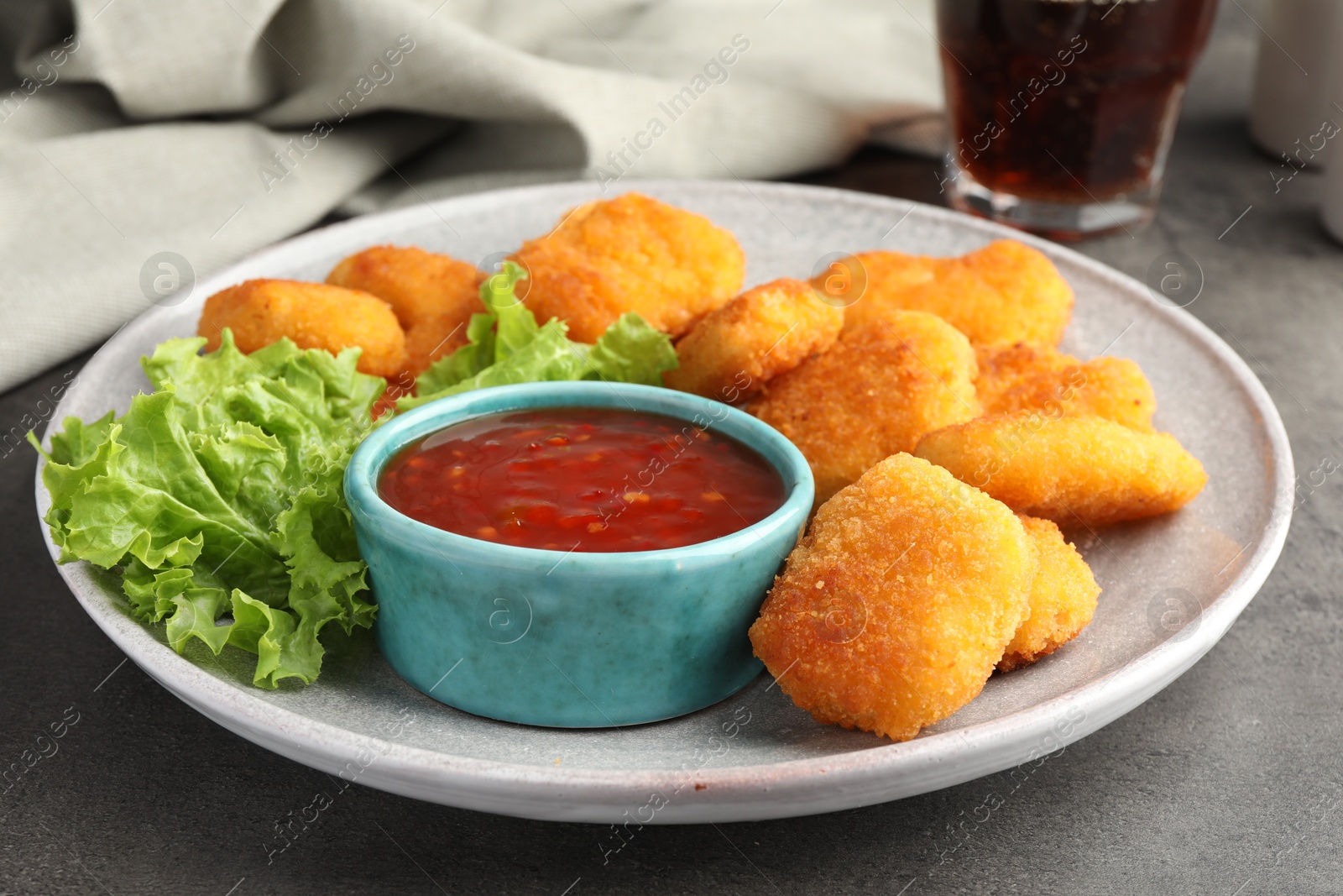 Photo of Plate with hot chili sauce, lettuce and nuggets on grey textured table, closeup