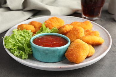 Photo of Plate with hot chili sauce, lettuce and nuggets on grey textured table, closeup