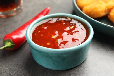 Photo of Hot chili sauce in bowl and pepper on grey textured table, closeup