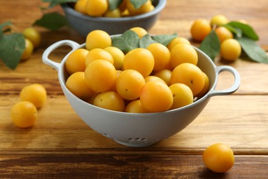 Photo of Tasty ripe plums and leaves in colander on wooden table, closeup