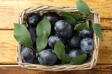 Photo of Tasty ripe plums and leaves in wicker basket on wooden table, top view