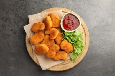 Photo of Board with hot chili sauce, lettuce and nuggets on grey textured table, top view