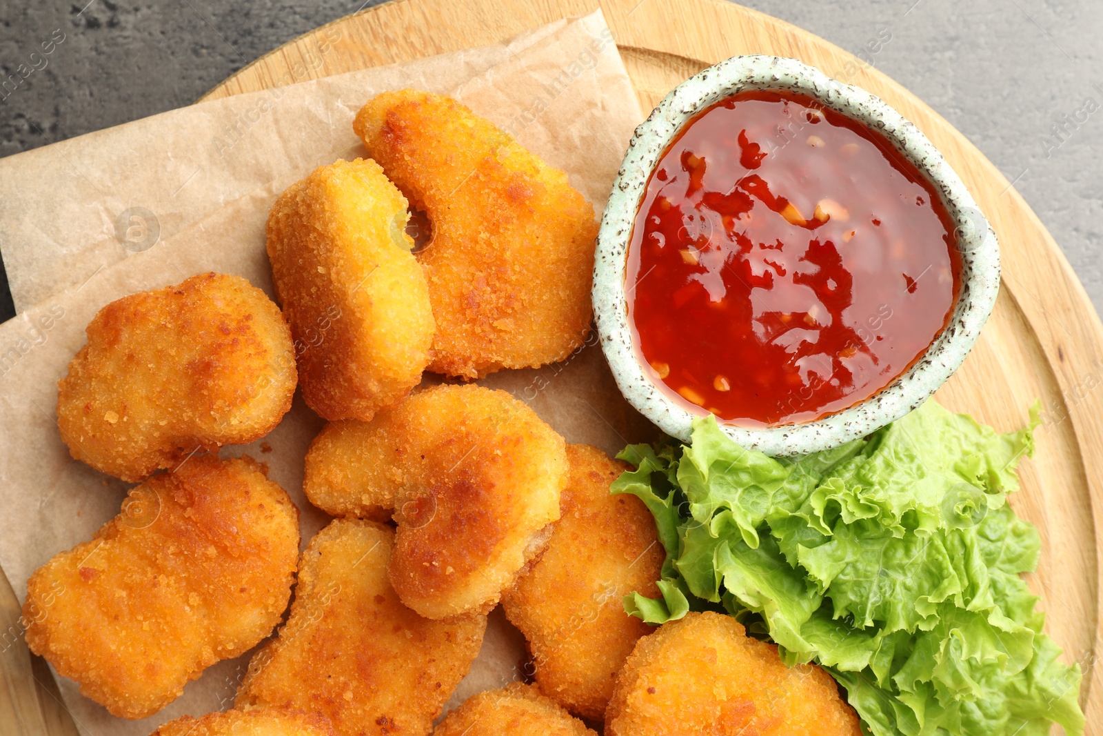 Photo of Board with hot chili sauce, lettuce and nuggets on grey textured table, top view