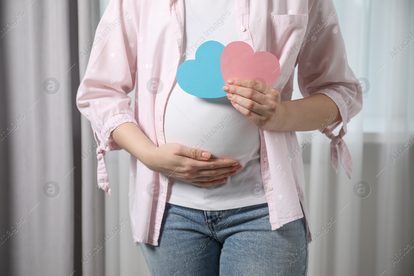 Photo of Expecting twins. Pregnant woman holding two paper cutouts of hearts at home, closeup