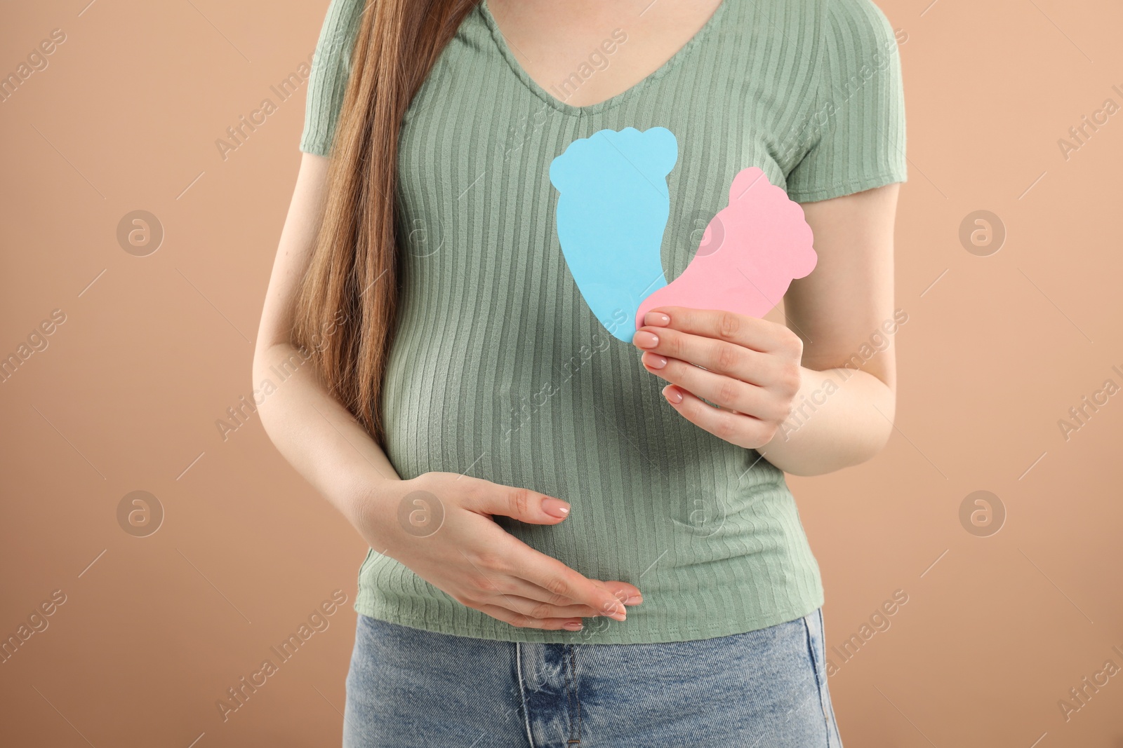 Photo of Expecting twins. Pregnant woman holding two paper cutouts of feet on light brown background, closeup