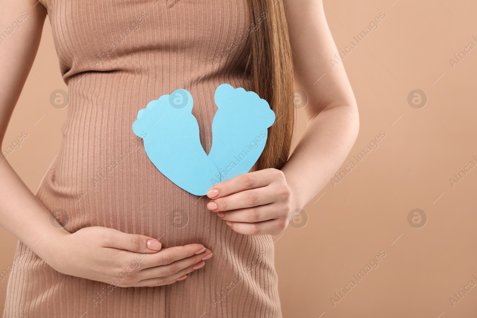 Photo of Expecting twins. Pregnant woman holding two paper cutouts of feet on light brown background, closeup. Space for text