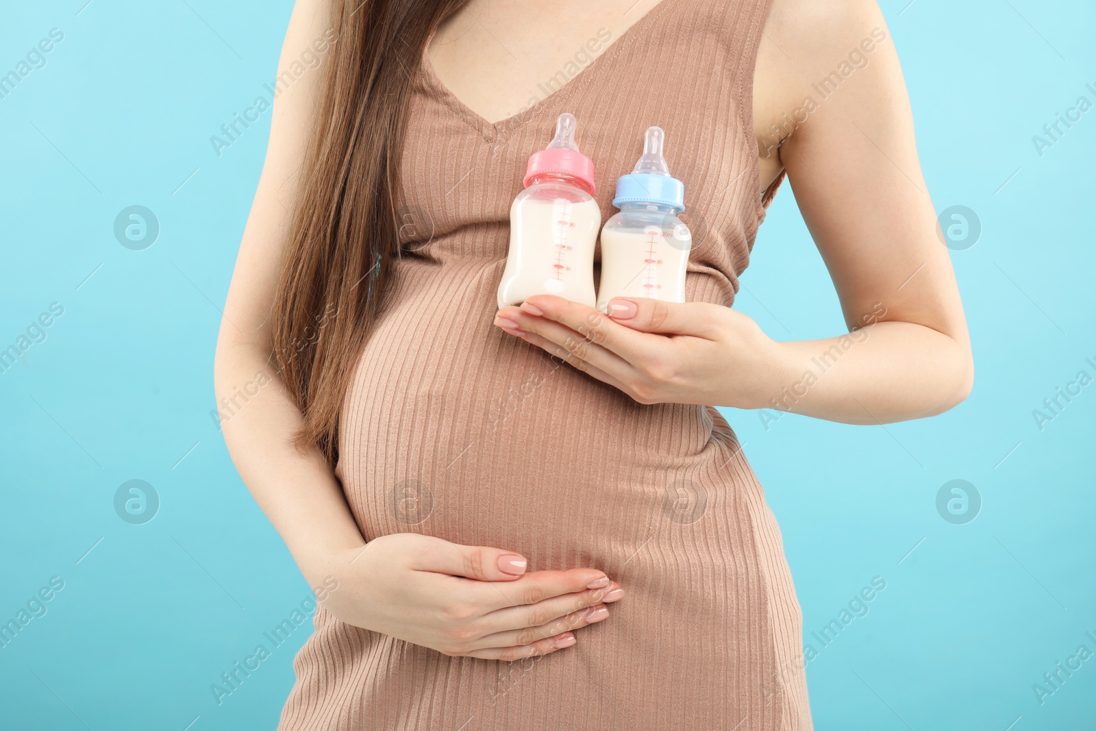 Photo of Expecting twins. Pregnant woman holding two bottles with milk on light blue background, closeup