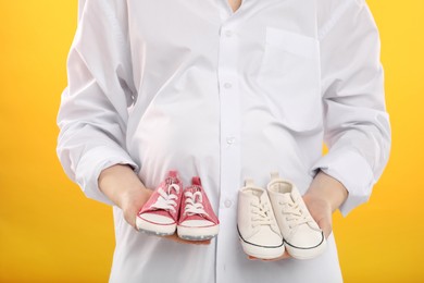 Expecting twins. Pregnant woman holding two pairs of shoes on yellow background, closeup