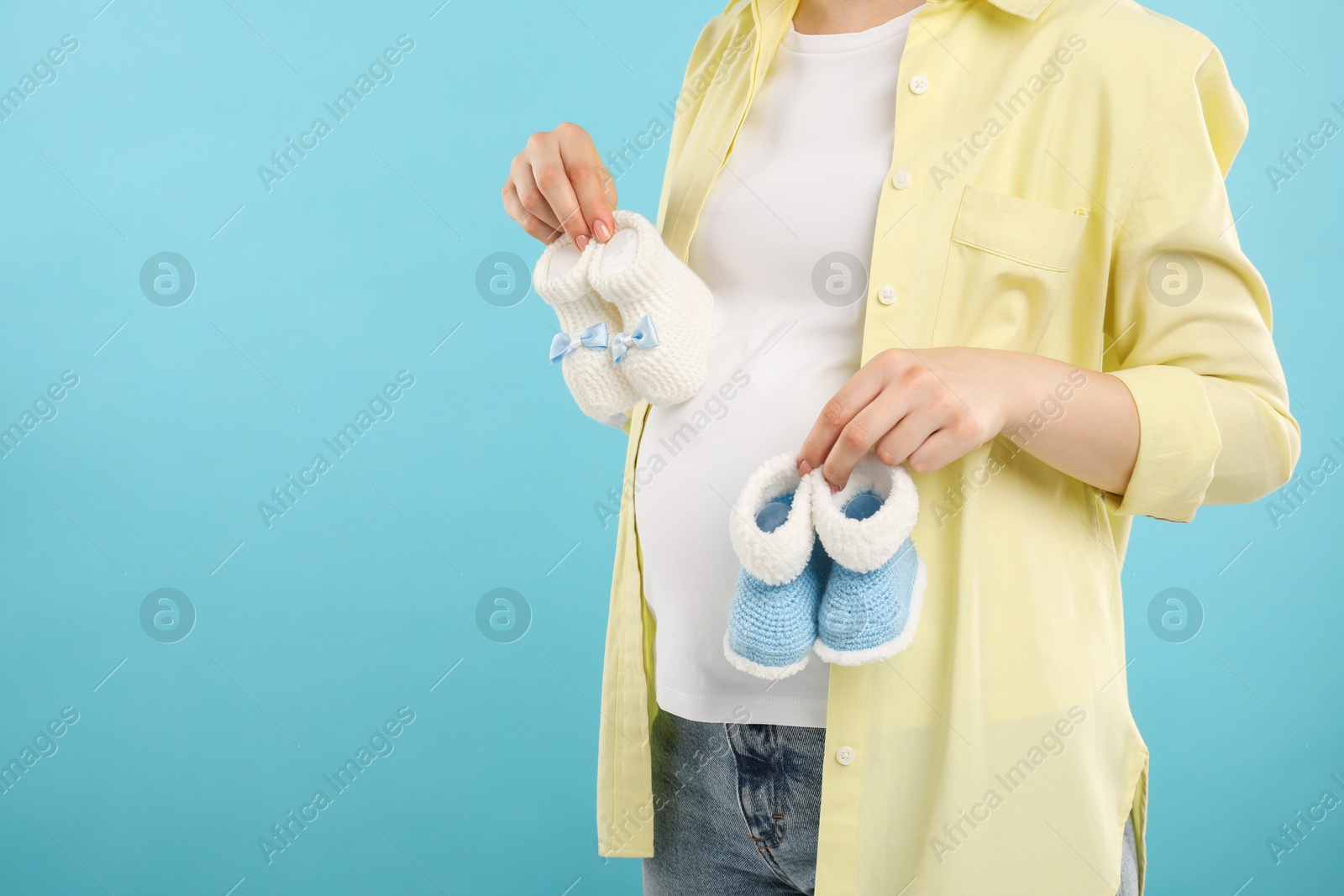 Photo of Expecting twins. Pregnant woman holding two pairs of baby shoes on light blue background, closeup. Space for text