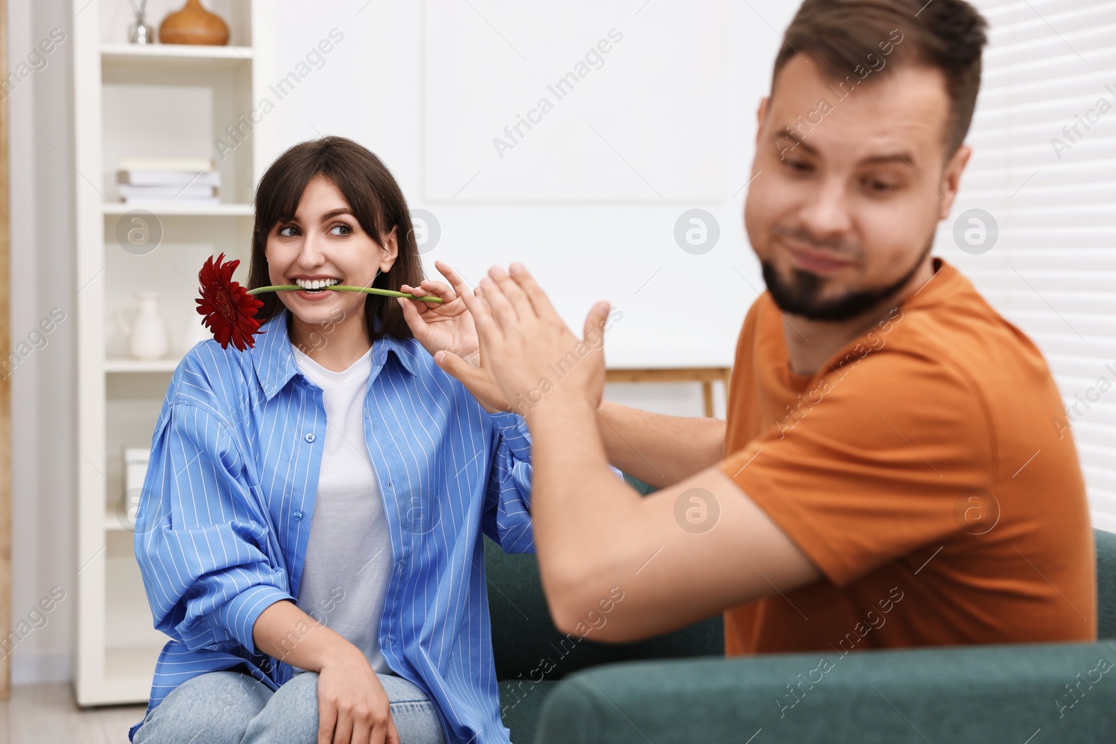 Photo of Woman with flower in mouth flirting with embarrassed man at home, selective focus