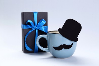 Photo of Happy Father's Day. Cup of coffee with funny paper hat, moustache and gift on light background