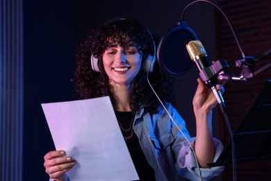 Photo of Woman with headphones singing into microphone in professional record studio