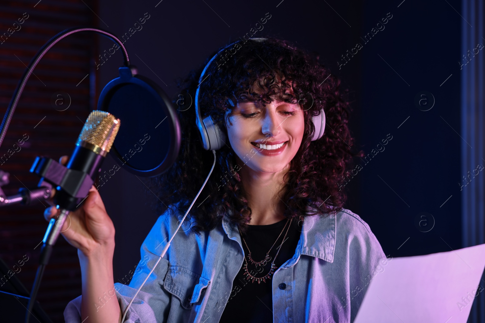 Photo of Woman with headphones singing into microphone in professional record studio