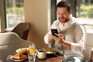 Photo of Happy man having tasty breakfast and using smartphone in cafe