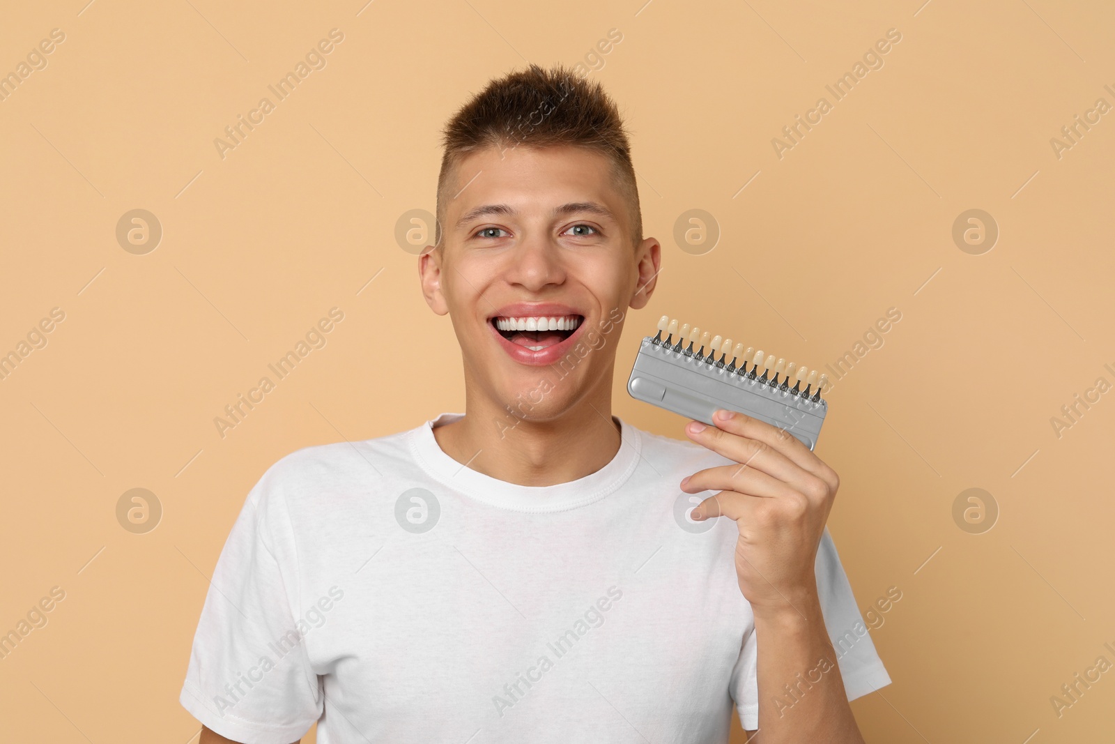 Photo of Happy young man with teeth color samples on beige background. Dental veneers