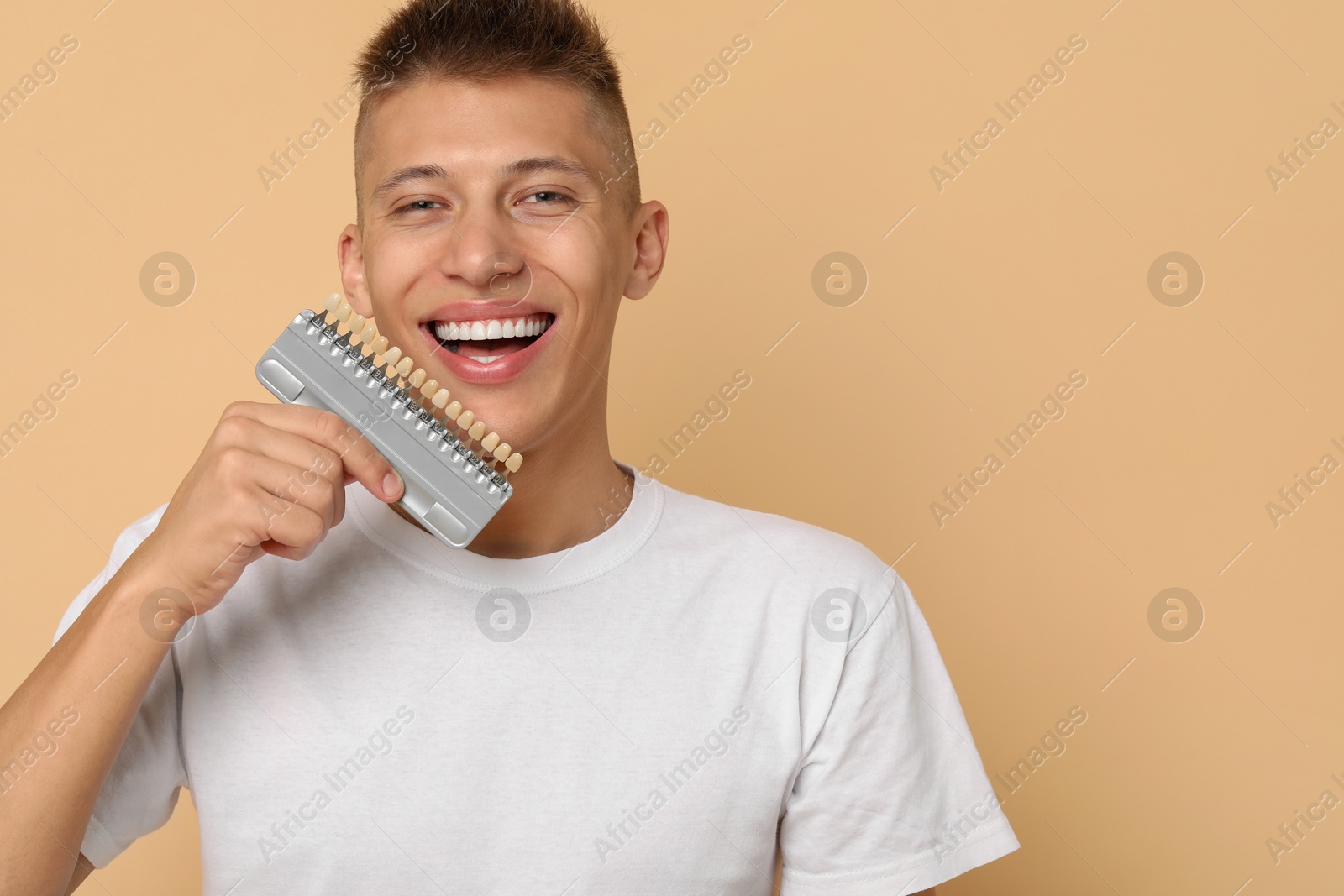 Photo of Happy young man with teeth color samples on beige background, space for text. Dental veneers