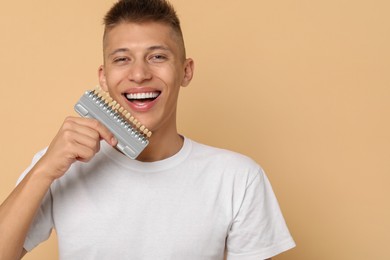 Photo of Happy young man with teeth color samples on beige background, space for text. Dental veneers