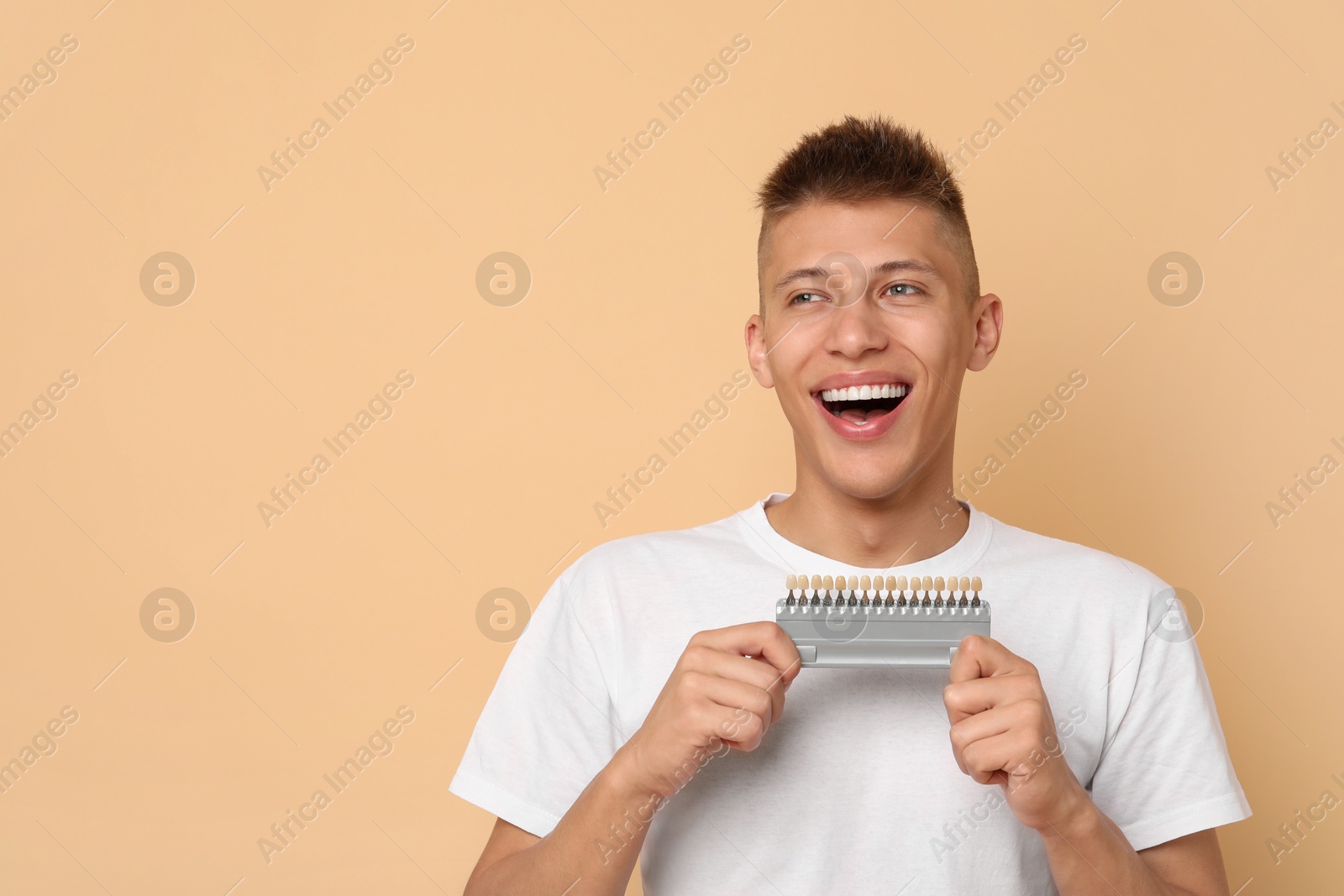 Photo of Happy young man with teeth color samples on beige background, space for text. Dental veneers