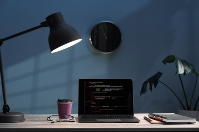 Photo of Programmer's workplace. Modern laptop with unfinished code, lamp and other supplies on wooden desk indoors