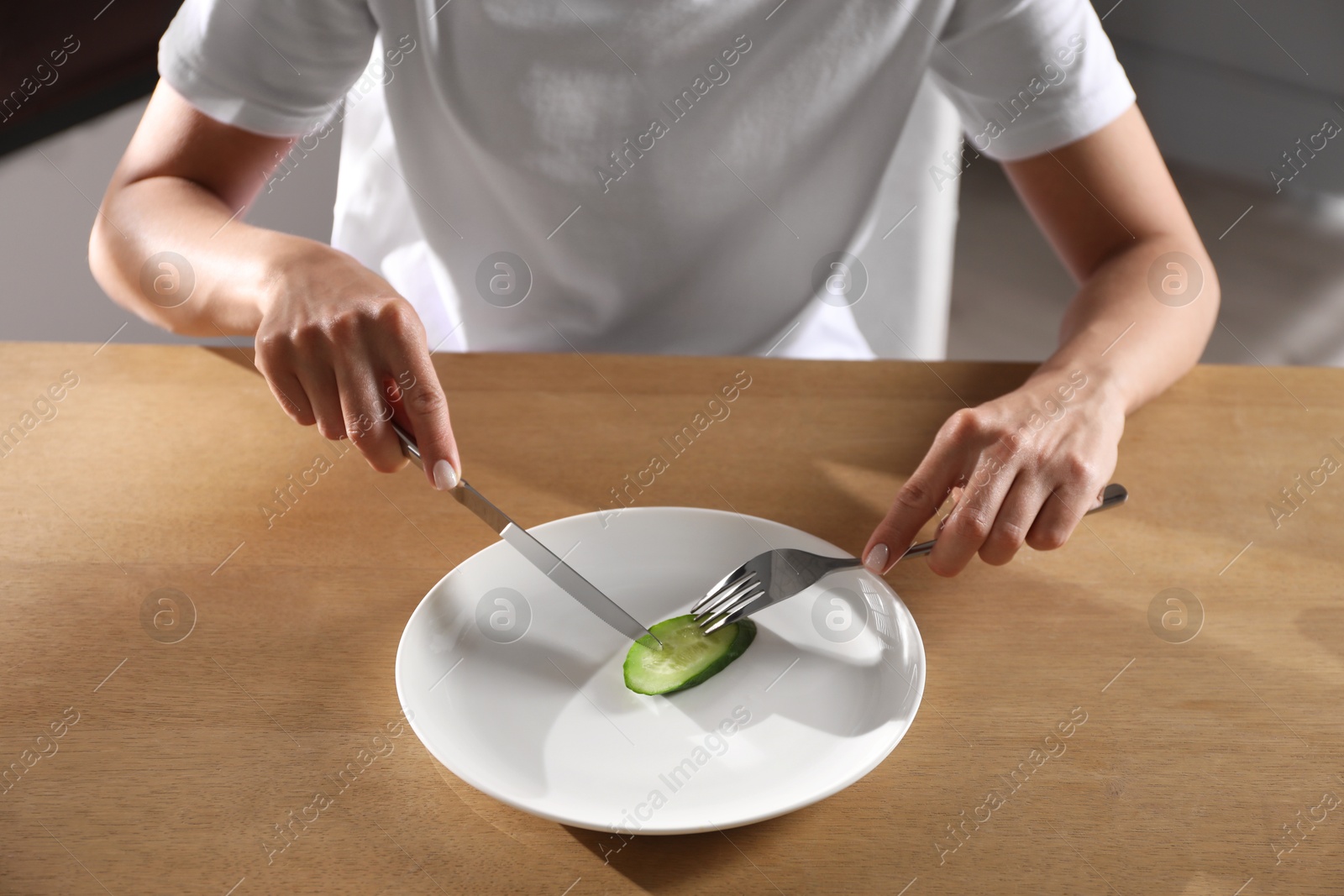 Photo of Eating disorder. Woman cutting cucumber at wooden table indoors, closeup