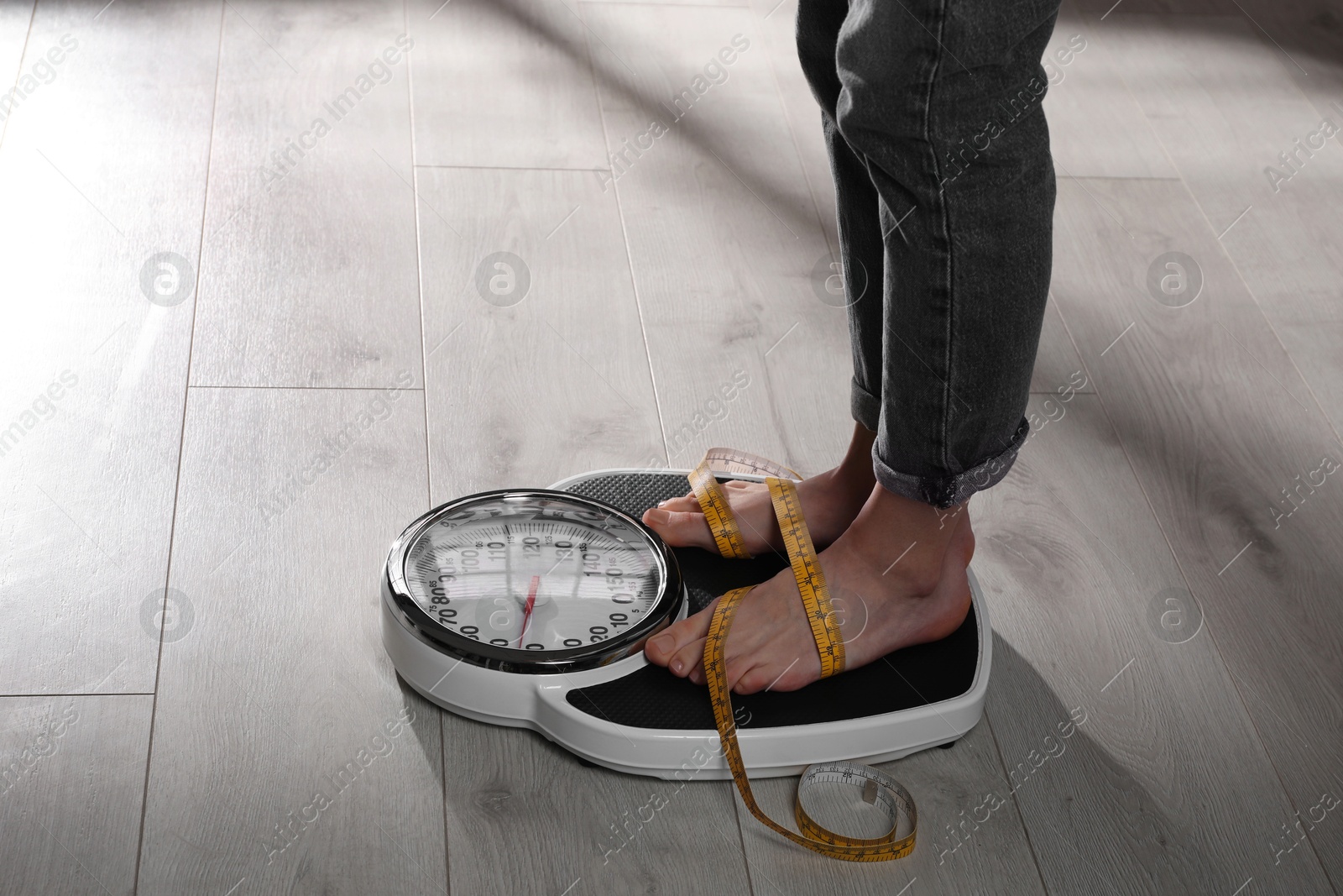 Photo of Eating disorder. Woman tied with measuring tape standing on floor scale indoors, closeup