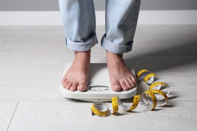 Photo of Eating disorder. Woman standing on floor scale and measuring tape indoors, closeup