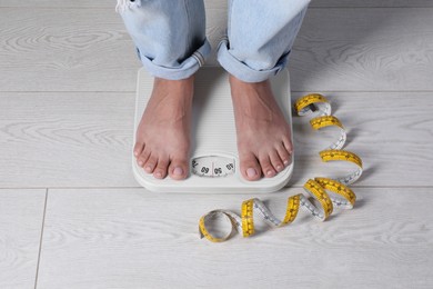 Photo of Eating disorder. Woman standing on floor scale and measuring tape indoors, closeup