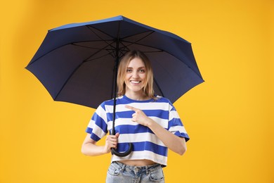 Woman with blue umbrella pointing at something on yellow background