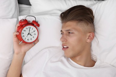 Sleepy young man in bed with alarm clock waking up at lunch time, top view