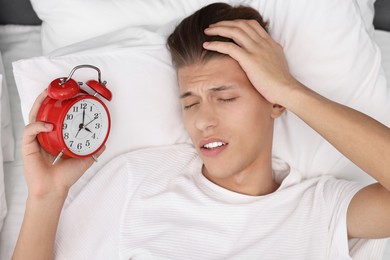 Sleepy young man in bed with alarm clock waking up at lunch time, top view