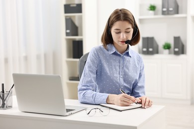 Teenager in headset taking notes while working at table indoors. Remote job