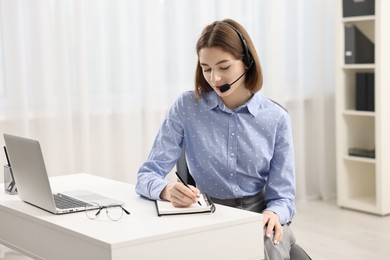 Teenager in headset taking notes while working at table indoors. Remote job