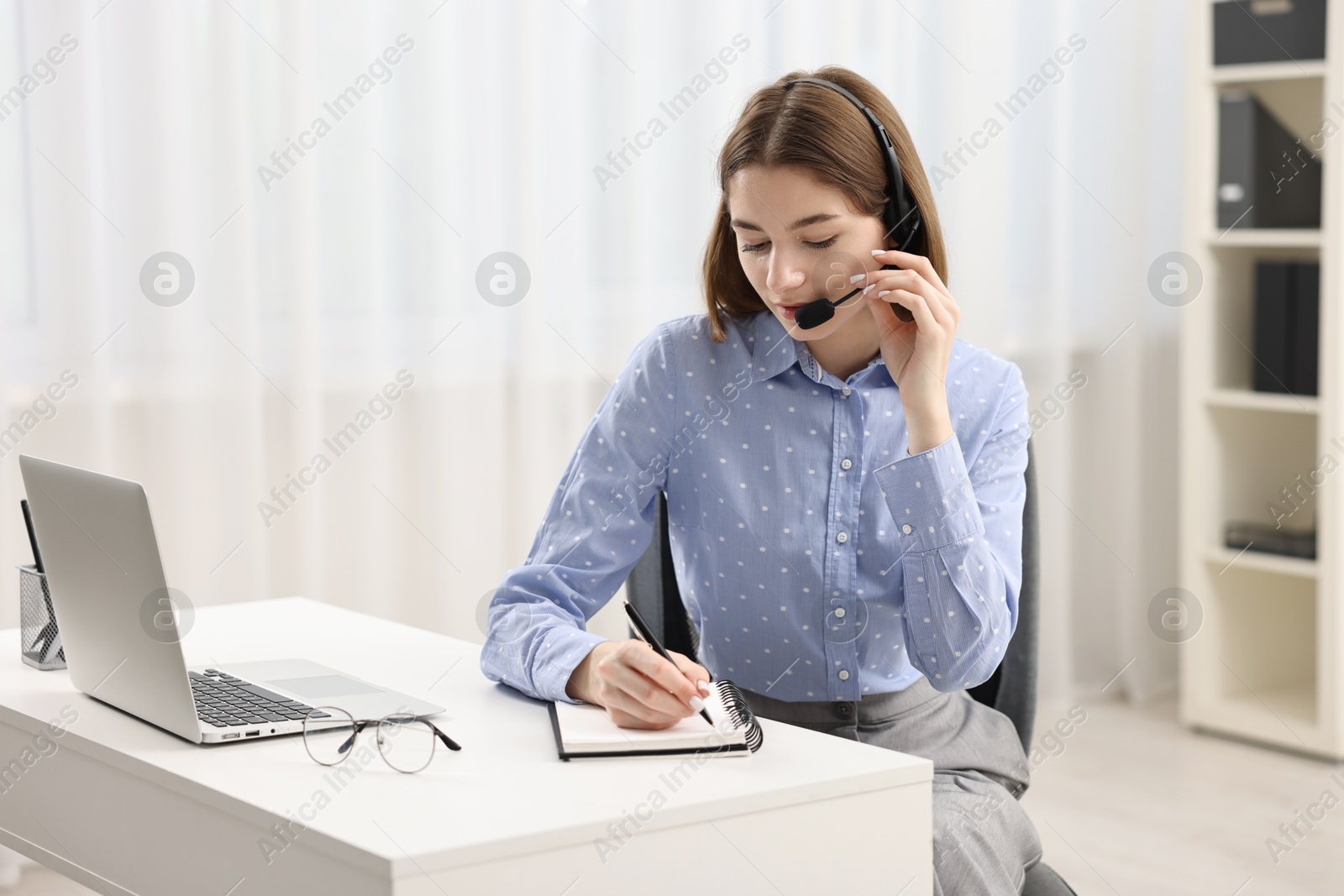 Photo of Teenager in headset taking notes while working at table indoors. Remote job