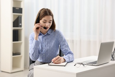 Teenager in headset taking notes while working at table indoors. Remote job